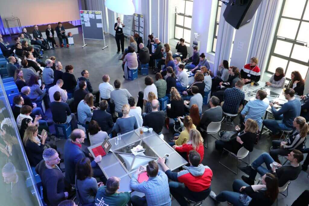 Bild zeigt eine Menschenmenge bei einem BarCamp. Die Moderatorin Ute Blindert steht vor dem Sessionplan.