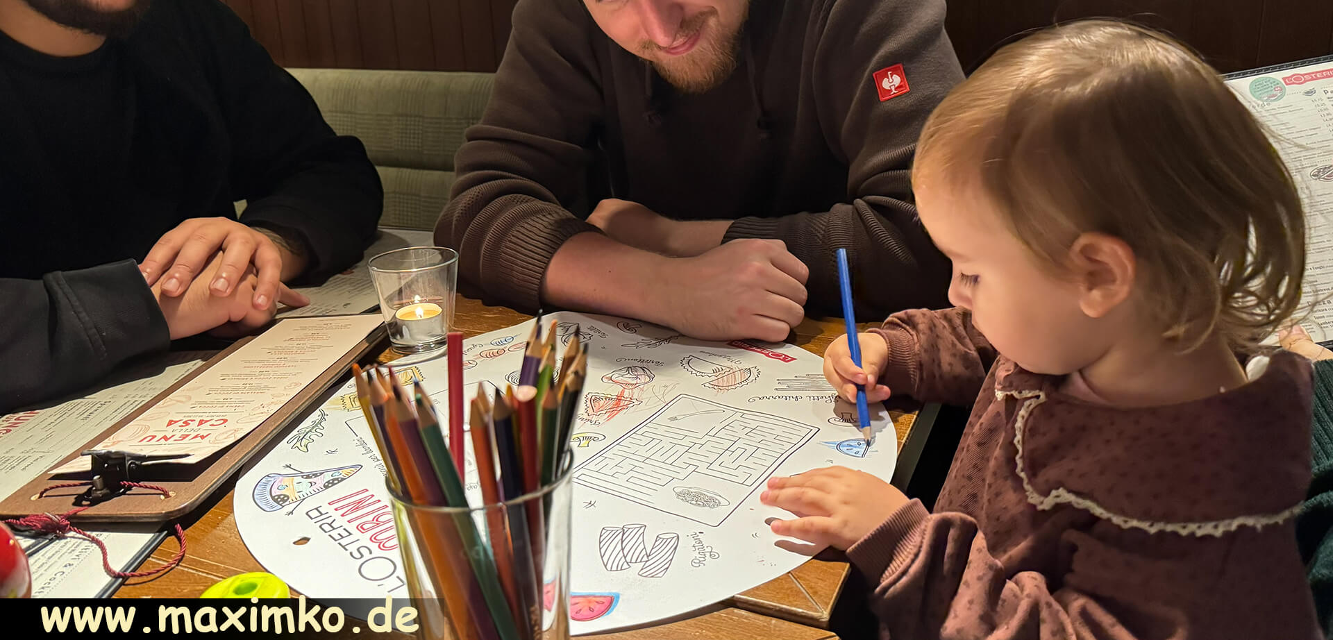 Zweijähriges Kind zeichnet mit Malstiften an einem Restauranttisch, während die Familie am Tisch zuschaut.
