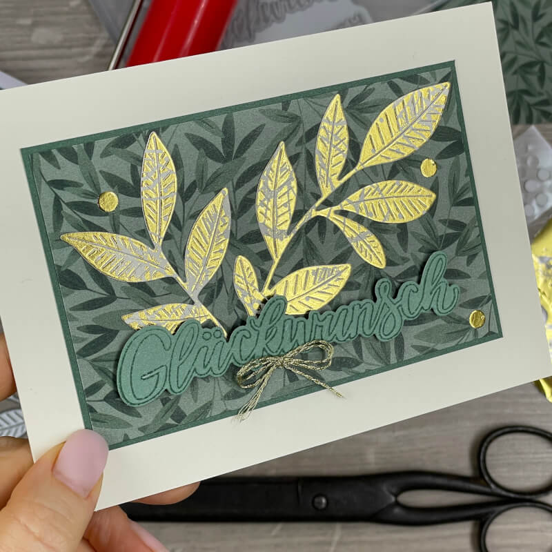 Einfachere Kartenversion mit grünem Designerpapier, goldfolierten Blattmotiven und Schriftzug 'Glückwunsch', in der Hand gehalten.