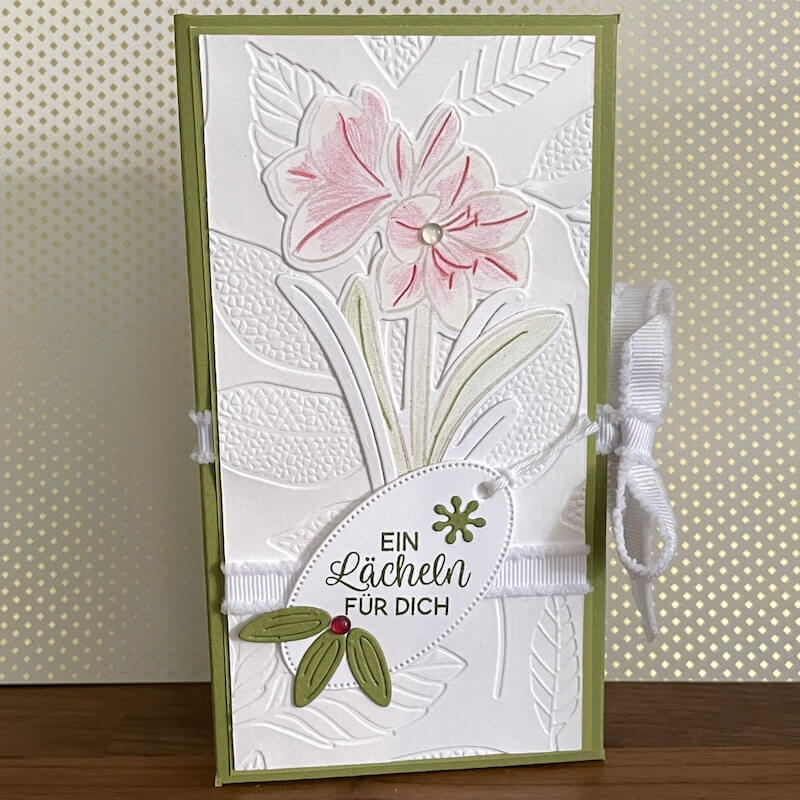 Zweite Deko-Variante mit zwei rosa Amaryllis-Blüten, geprägtem Blattmuster und weißer Satinschleife auf Waldmoos