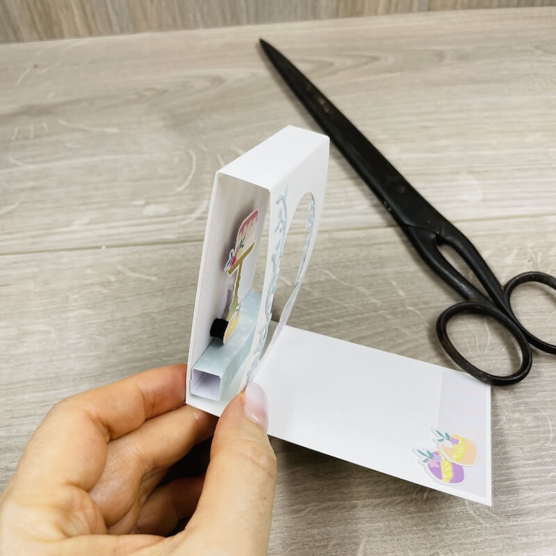 Karte 11 von der Seite: die aufgestellte Mini-Aufstellkarte in der Hand, dahinter eine Schere