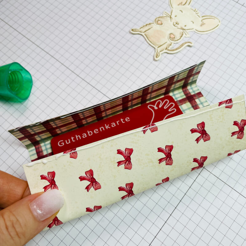 Designerpapier mit roten Schleifen liegt locker um eine Gutscheinkarte, die Ränder sind mit Kleber eingestrichen