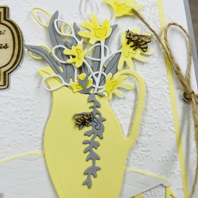 Nahaufnahme des gelben Krugs mit Blütenstrauß, gelasertem Bienchen und der Juteschnur, die die Karte hält