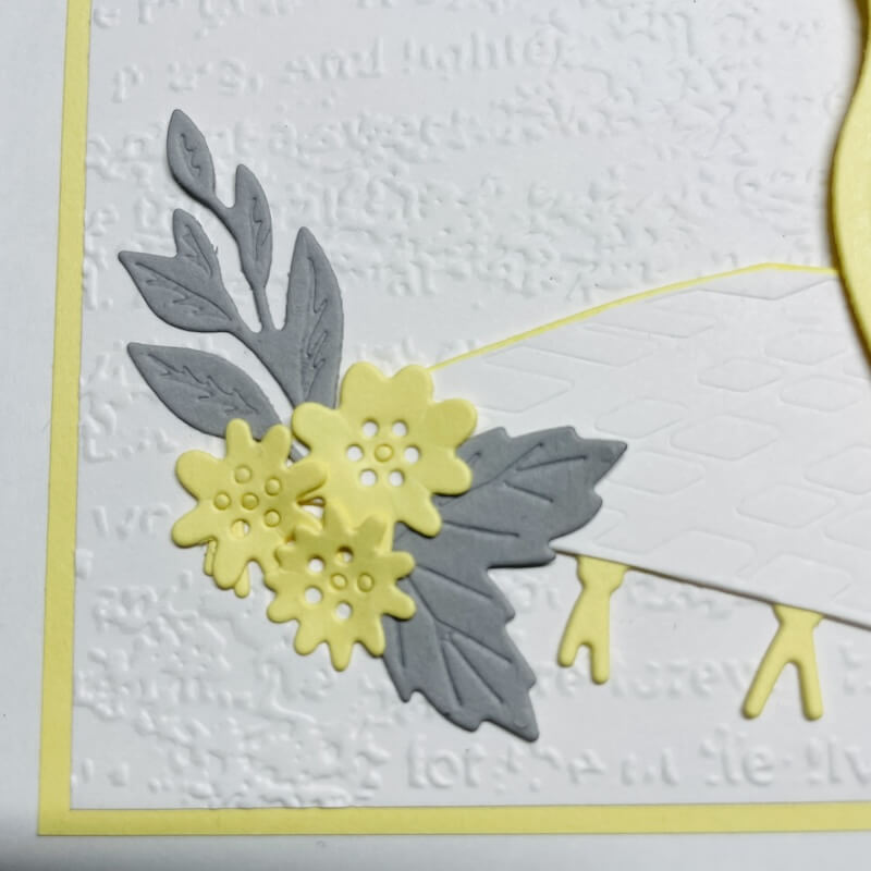 Detail der Kartenvorderseite: gelbe Blüten mit geprägter Mitte, graue Blätter und eine kleine Wimpelkette