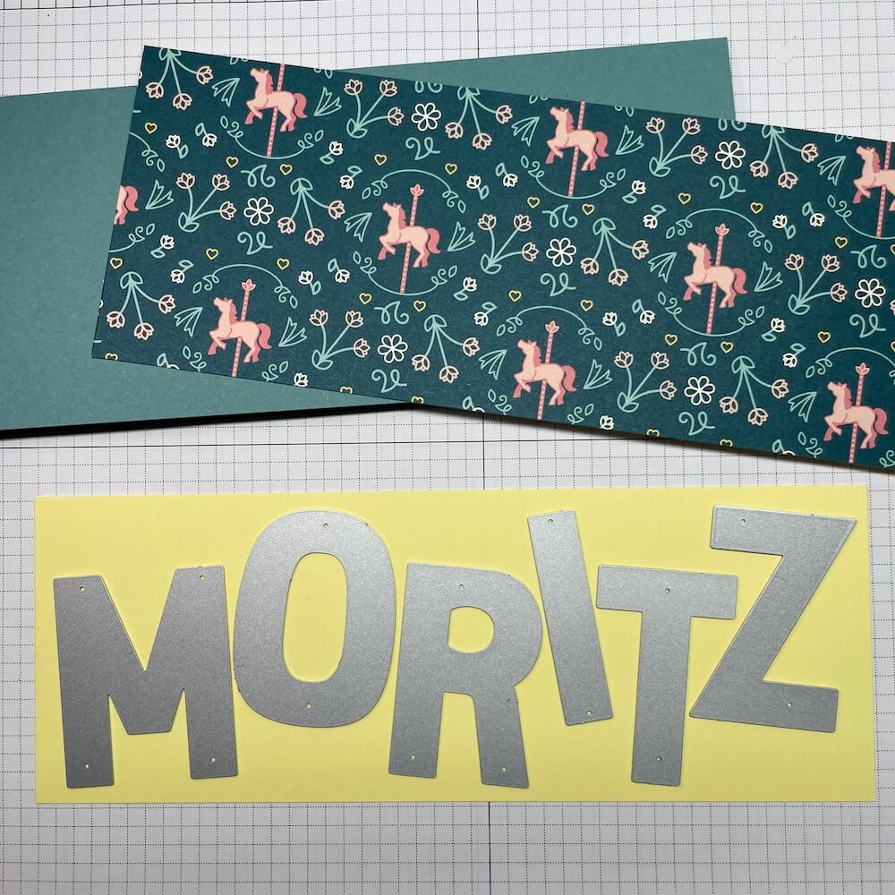 Buchstabenstanzen des Party-Alphabets bilden den Namen Moritz auf zitronengelbem Farbkarton, darüber Designerpapier und Lagunenblau