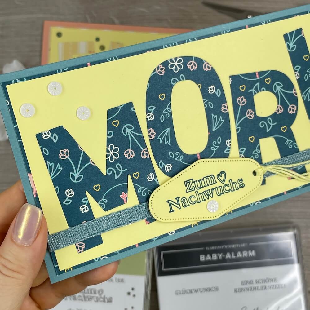 Karte für Moritz in der Hand: Buchstaben aus dunkelblauem Designerpapier auf zitronengelbem Aufleger mit Jeansband und Label Zum Nachwuchs