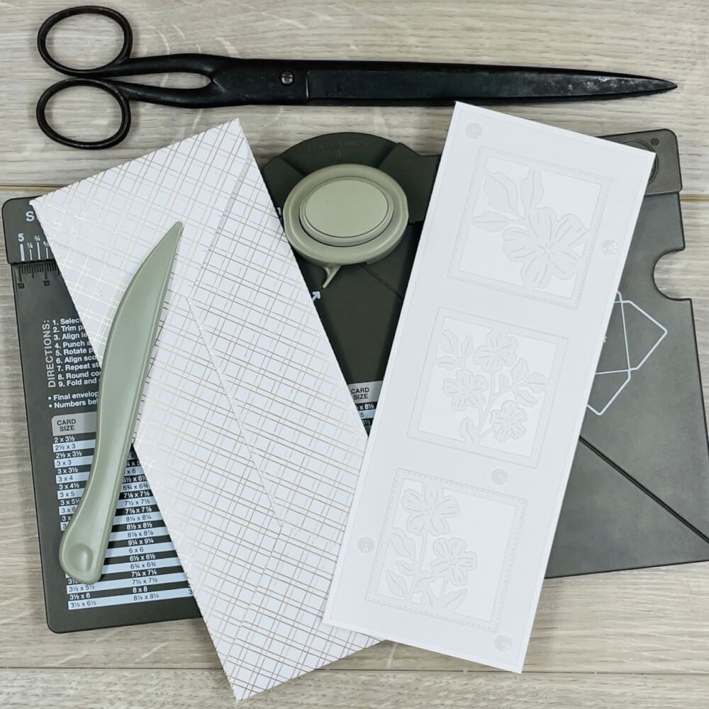 Schmale Karte mit geprägten Blüten liegt mit Falzbein und Papierschere auf dem Envelope Punch Board