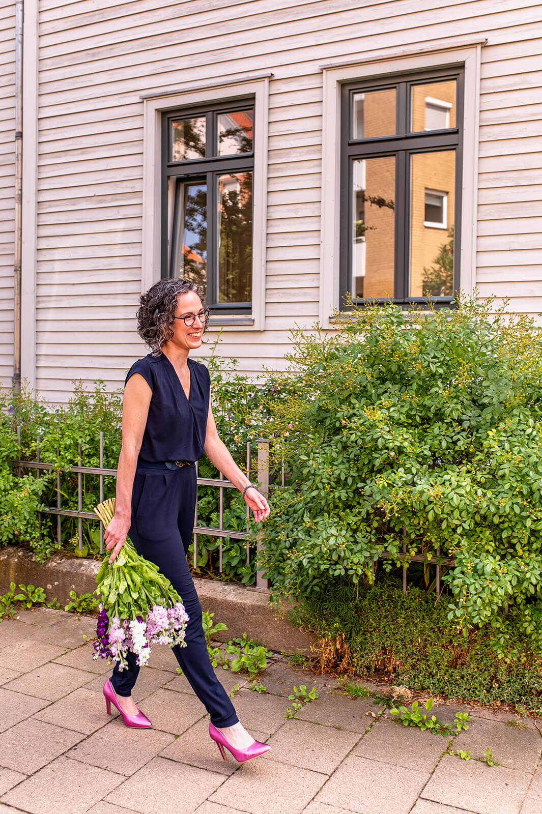 Annika Lind läuft mit Blumenstrauß an einer Hausfassade entlang, trägt einen dunklen Jumpsuit und pinke Pumps.