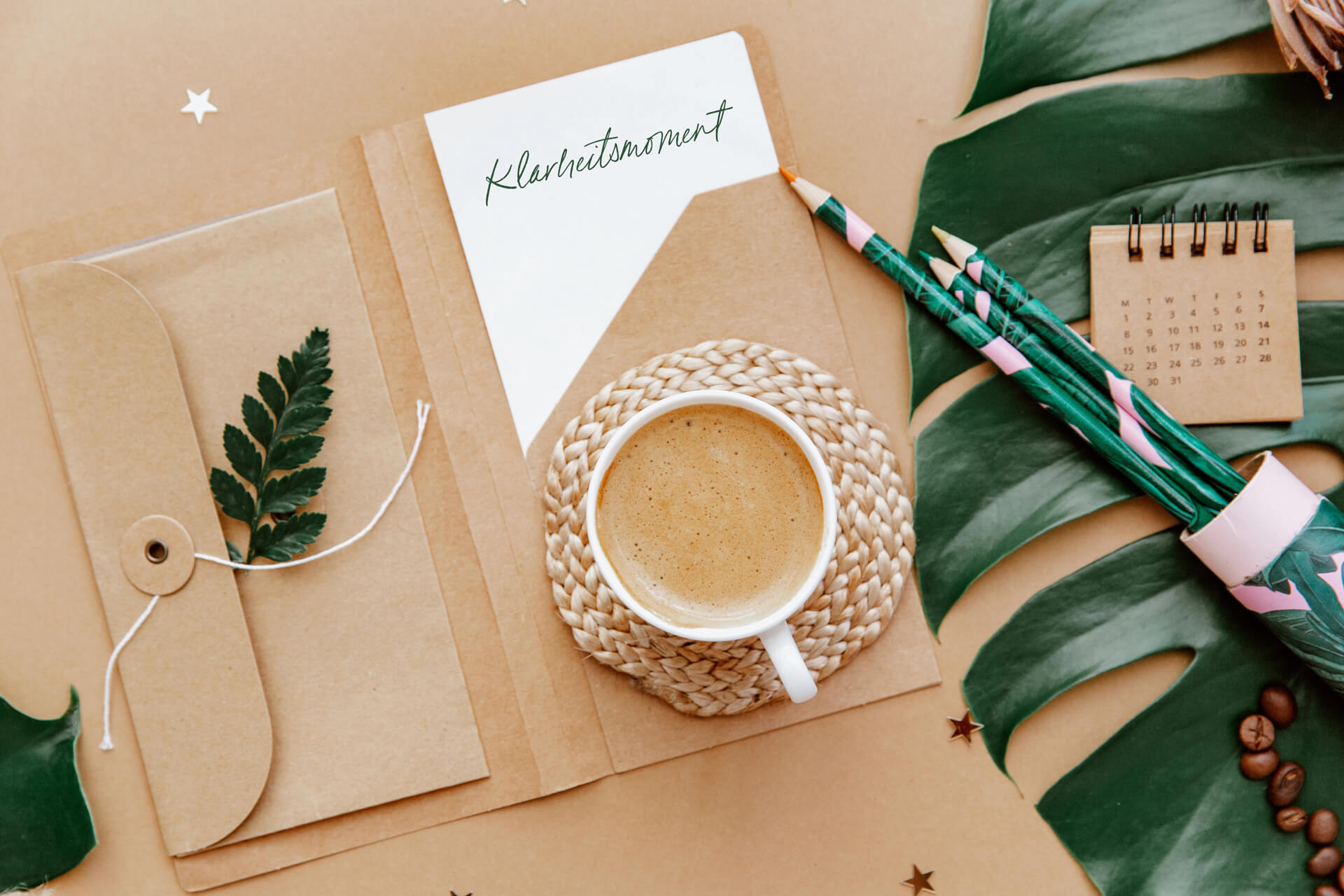 Beiges Flatlay mit Notizkarte, Umschlag, grünem Blatt, Kaffee und Bleistiften auf tropischem Hintergrund – Symbol für Ruhe, Fokus und kreative Klarheit im Branding-Prozess.