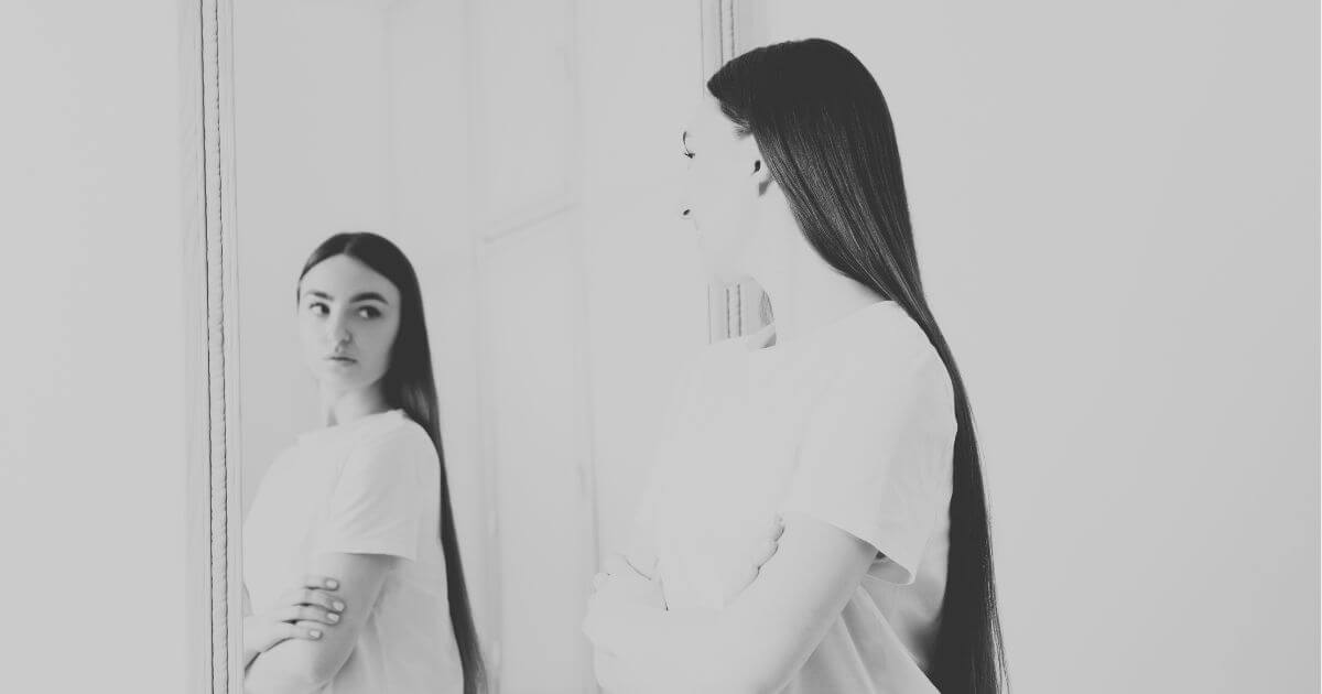 Junge Frau blickt nachdenklich in einen Spiegel – Sinnbild für inneren Kritiker und Selbstkritik.