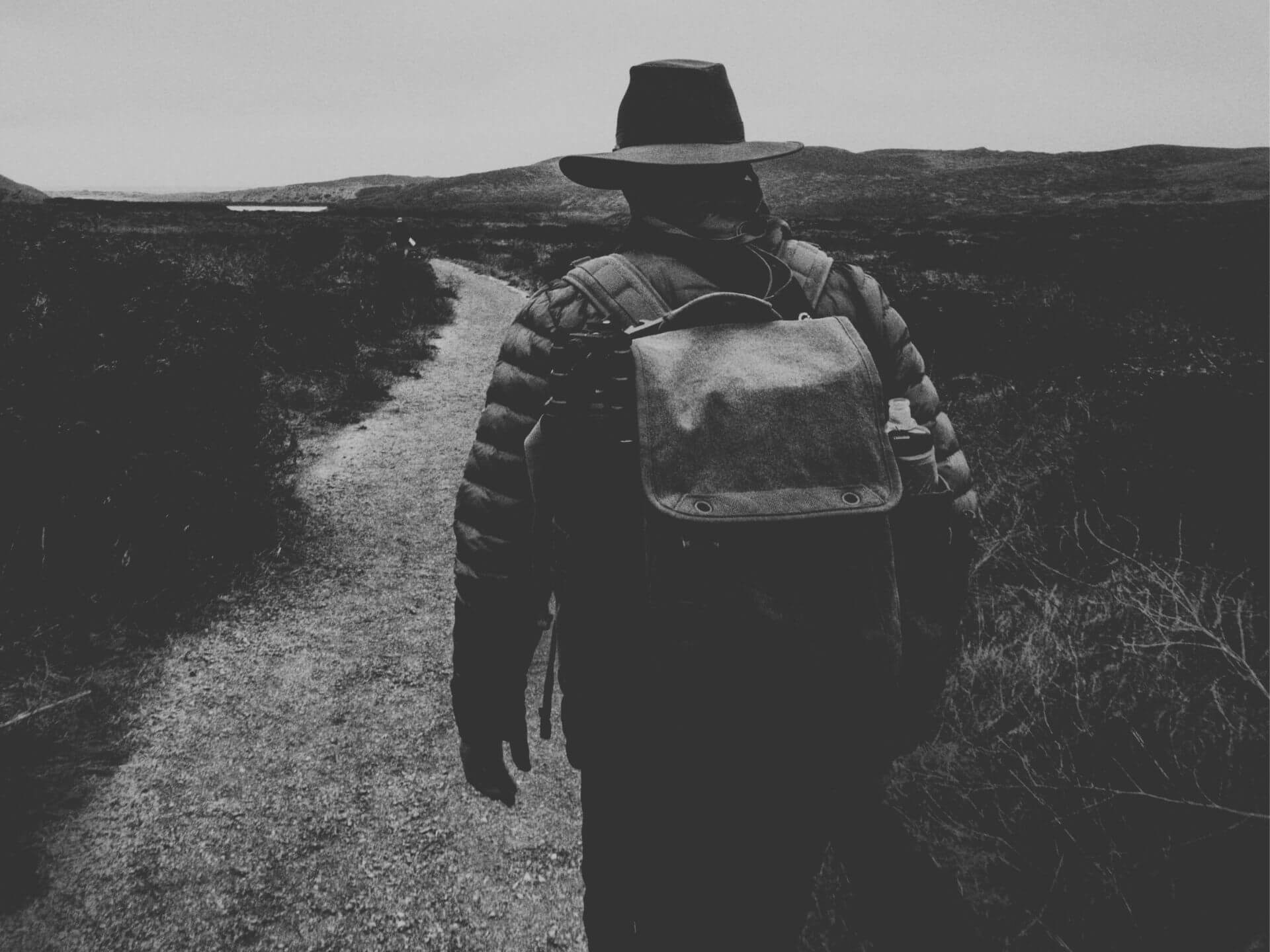 Person mit großem Rucksack auf einem Weg in einer offenen Landschaft – Sinnbild für die Summe von Belastungen, die zu Überforderung führen können.