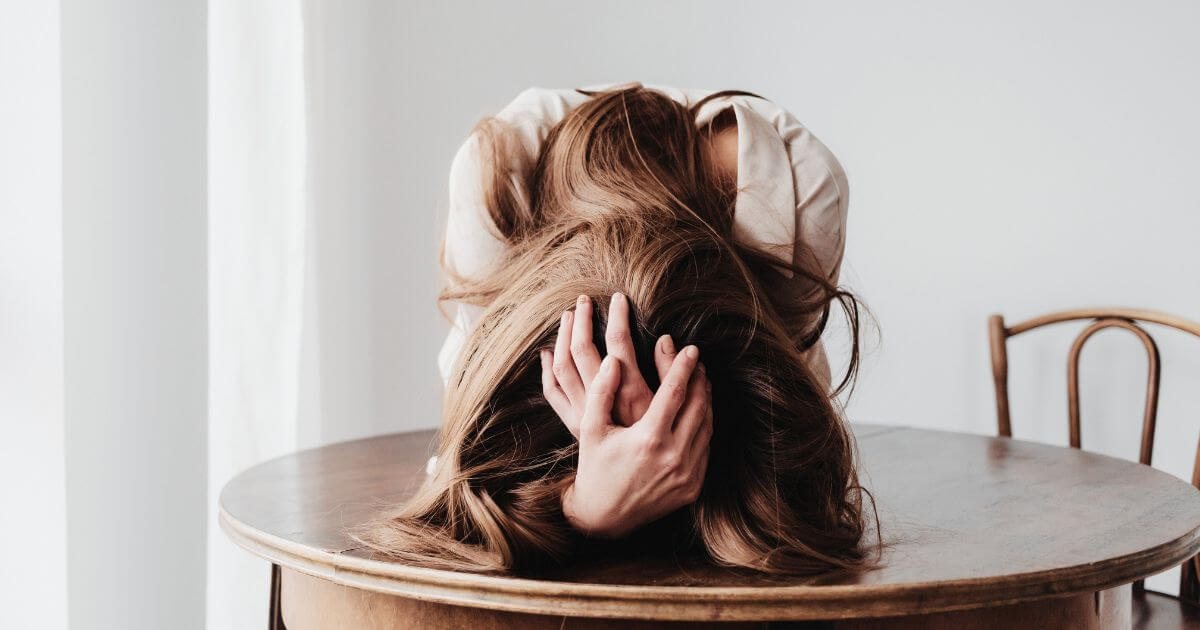 Eine Person sitzt erschöpft mit gesenktem Kopf an einem Tisch – symbolisches Bild für tiefe Erschöpfung, innere Überforderung und die körperlichen und psychischen Symptome von Erschöpfung.