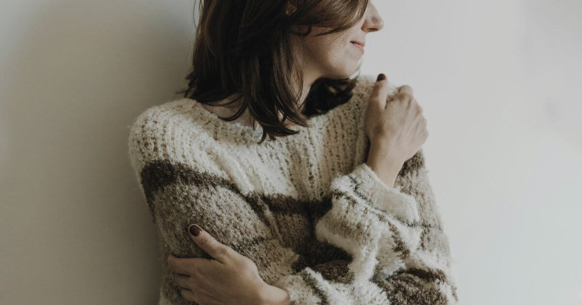 Person steht am Fenster und hält sich selbst – symbolisches Bild für inneren Kritiker, Selbstkritik und den Versuch von Selbstzuwendung.