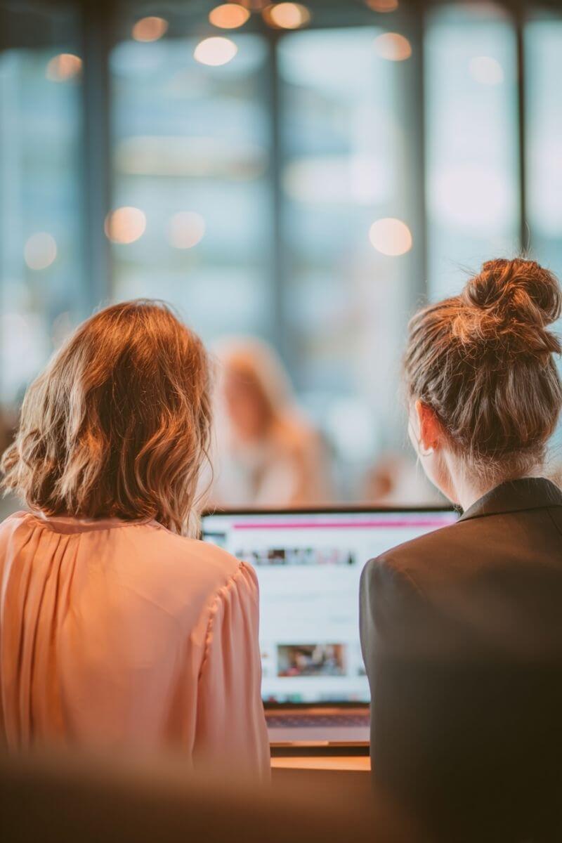 Zwei Frauen arbeiten gemeinsam an einem Laptop in einem modernen Büro. Eine Frau mit schulterlangem dunkelblondem Haar in roséfarbener Bluse sitzt neben einer Kollegin mit dunklem Dutt. Der Bildschirm zeigt ein soziales Netzwerk.