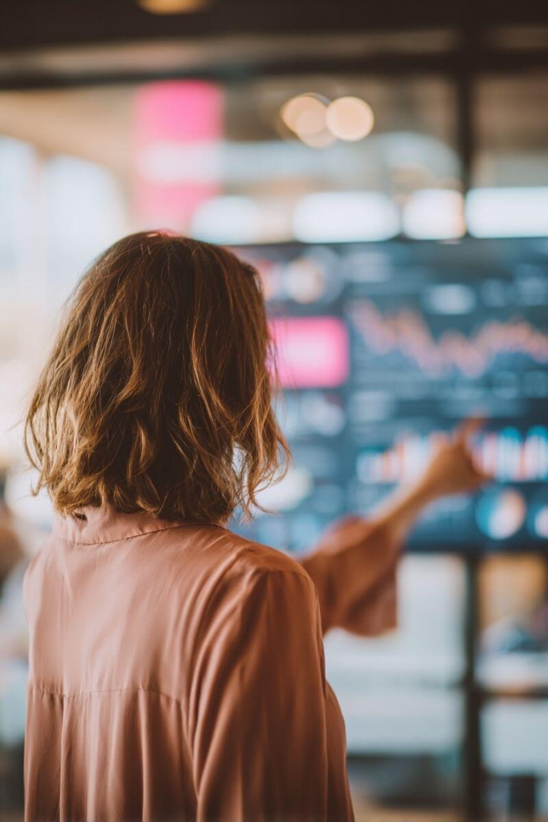 Online-Marketing-Expertin mit schulterlangem dunkelblondem Haar in roséfarbener Bluse zeigt in einem modernen Büro auf einen großen Monitor mit Diagrammen und Datenvisualisierungen.