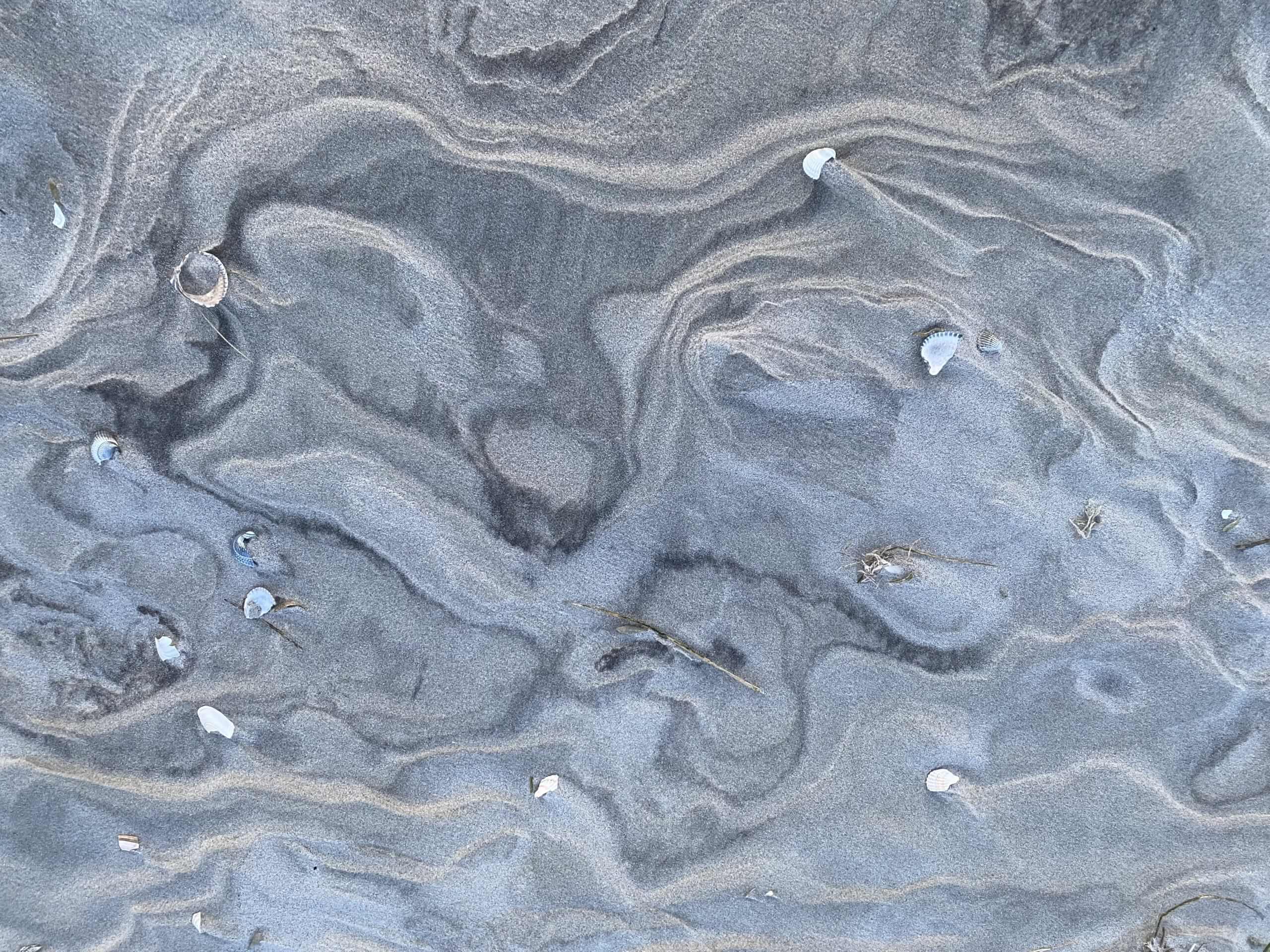 Strukturmuster im feuchten Sand am Strand, mit wellenförmigen Linien und kleinen Vertiefungen